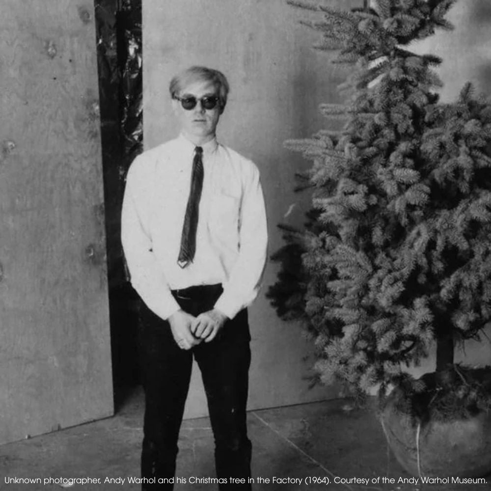 Black and white photo of Andy Warhol standing next to a Christmas tree in the Factory, 1964.