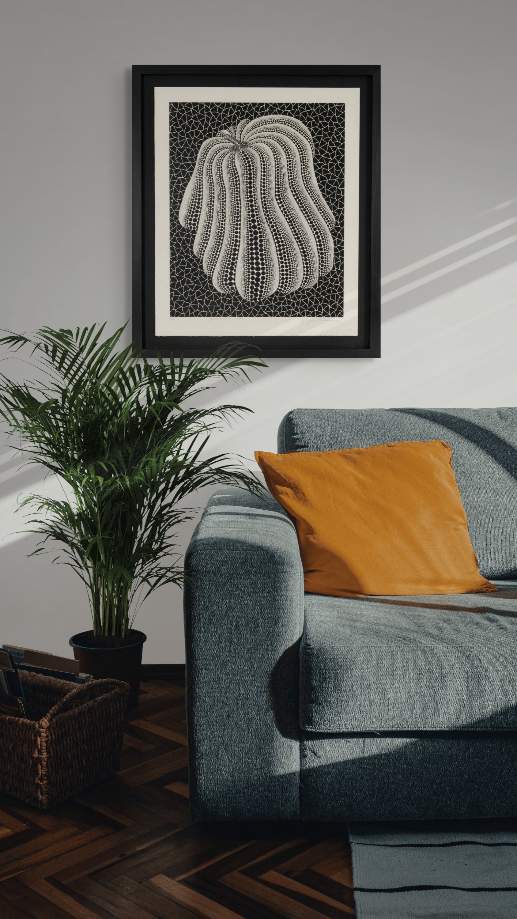 A framed black and white abstract Yayoi Kusama pumpkin art print featuring a curved striped pattern, displayed on a gray wall above a blue-gray sofa with an orange pillow and next to a potted plant.